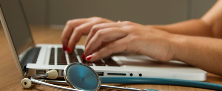 Person typing on a laptop with a stethoscope lying beside them on the desk.