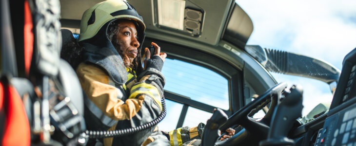 Side view of a Black female airport firefighter sitting in a fire truck using a CB station while driving towards an emergency location at the airport runway.