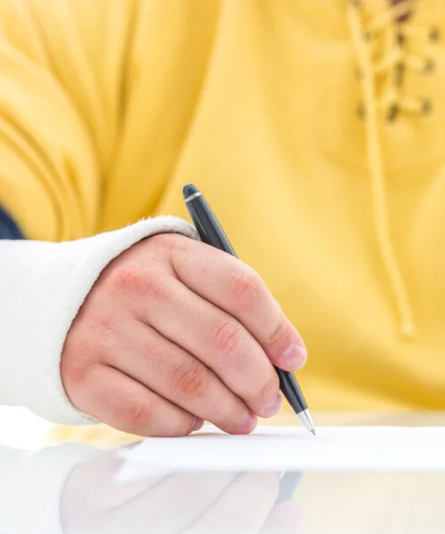 Close up of person wearing a yellow shirt with a cast on their hand signing a document